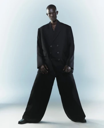 Fashion shot of a dark-skinned male model in an oversized black double-breasted suit and wide-leg pants against a gradient blue background.