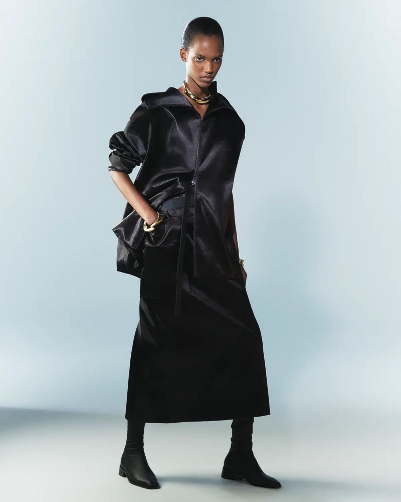 Fashion model in a black outfit posing in a studio with a light blue background, wearing gold jewelry and ankle boots.