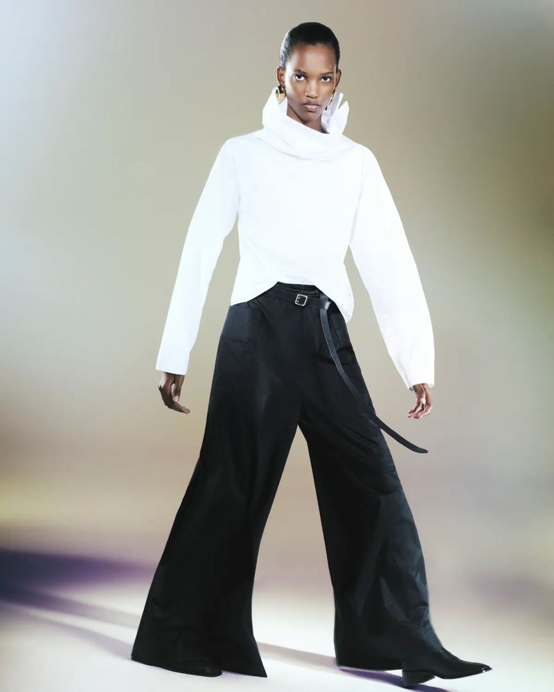 Fashion model in a studio wearing a white long-sleeved top and black wide-leg pants, posing with a neutral expression and soft lighting.