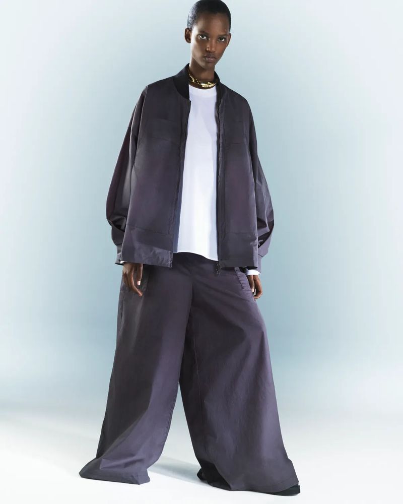 Model wearing a dark grey bomber jacket and matching wide-leg trousers over a white t-shirt against a light gradient background.