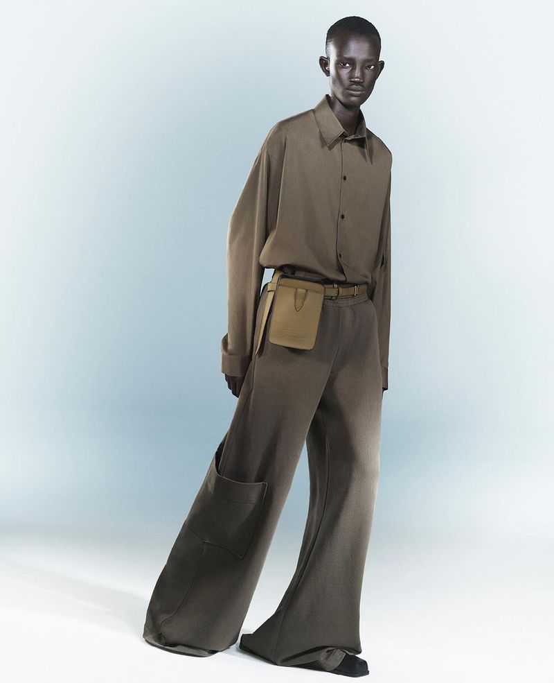 Fashion shot of a Black adult male model wearing a long-sleeved shirt and wide-leg pants with a small bag on a light blue background.