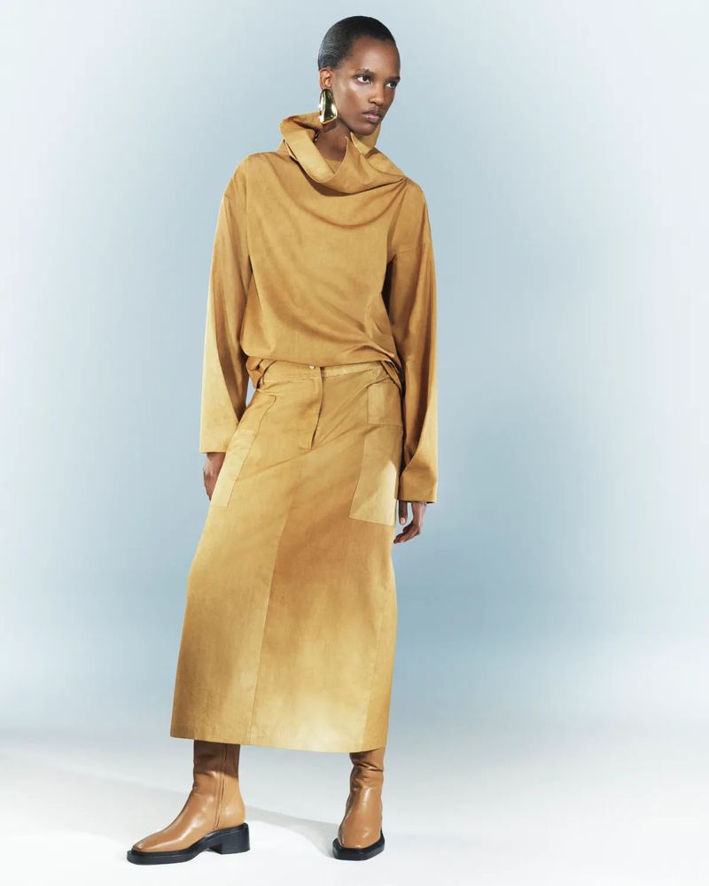 Woman in tan cowl-neck top and midi skirt poses in studio with neutral background, showcasing modern, minimalist, monochromatic fashion.