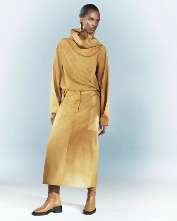 Woman in tan cowl-neck top and midi skirt poses in studio with neutral background, showcasing modern, minimalist, monochromatic fashion.