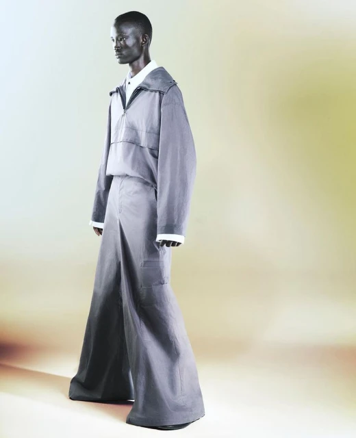An adult androgynous person stands in a photo studio wearing a gray oversized jacket and wide-leg pants with a yellow to brown gradient backdrop.