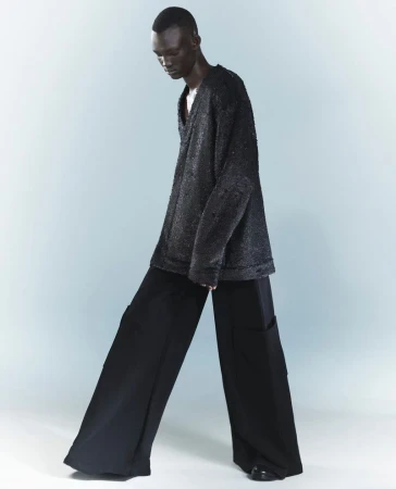 Model Goy Michael poses in an oversized dark textured knit sweater and ultra wide-leg black trousers against a pale studio backdrop.
