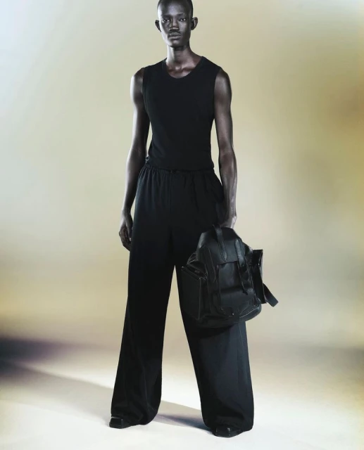 Man in all-black tank top and loose pants holding a backpack in a studio setting with soft lighting, fashionable and modern design.