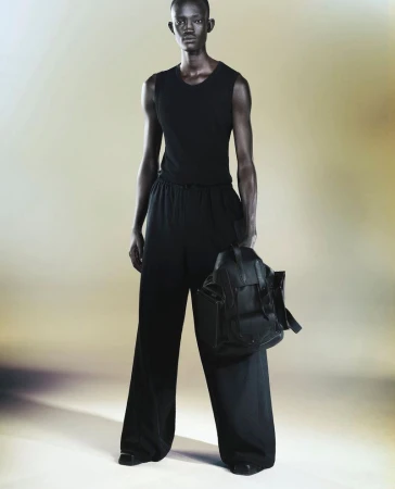 Man in all-black tank top and loose pants holding a backpack in a studio setting with soft lighting, fashionable and modern design.