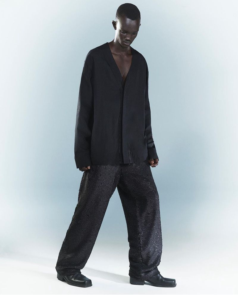 A dark-skinned adult model poses in a black V-neck top and patterned wide-leg trousers against a gradient grey-blue backdrop in a studio.
