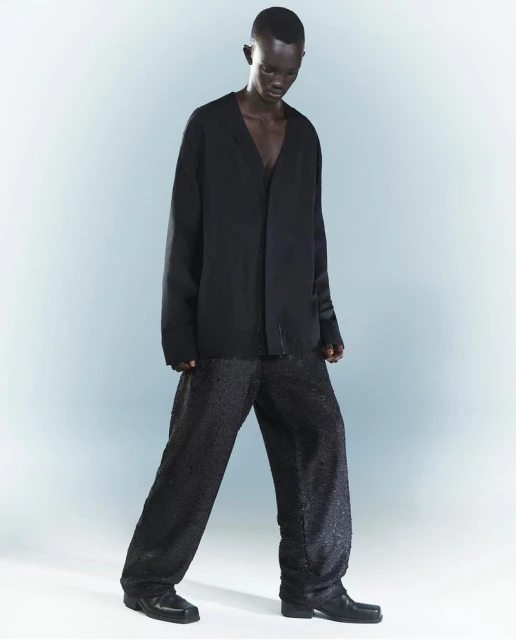 A dark-skinned adult model poses in a black V-neck top and patterned wide-leg trousers against a gradient grey-blue backdrop in a studio.