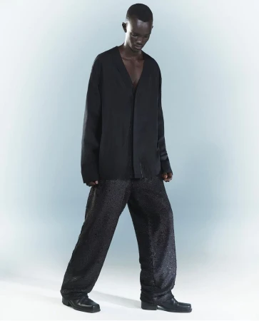 A dark-skinned adult model poses in a black V-neck top and patterned wide-leg trousers against a gradient grey-blue backdrop in a studio.