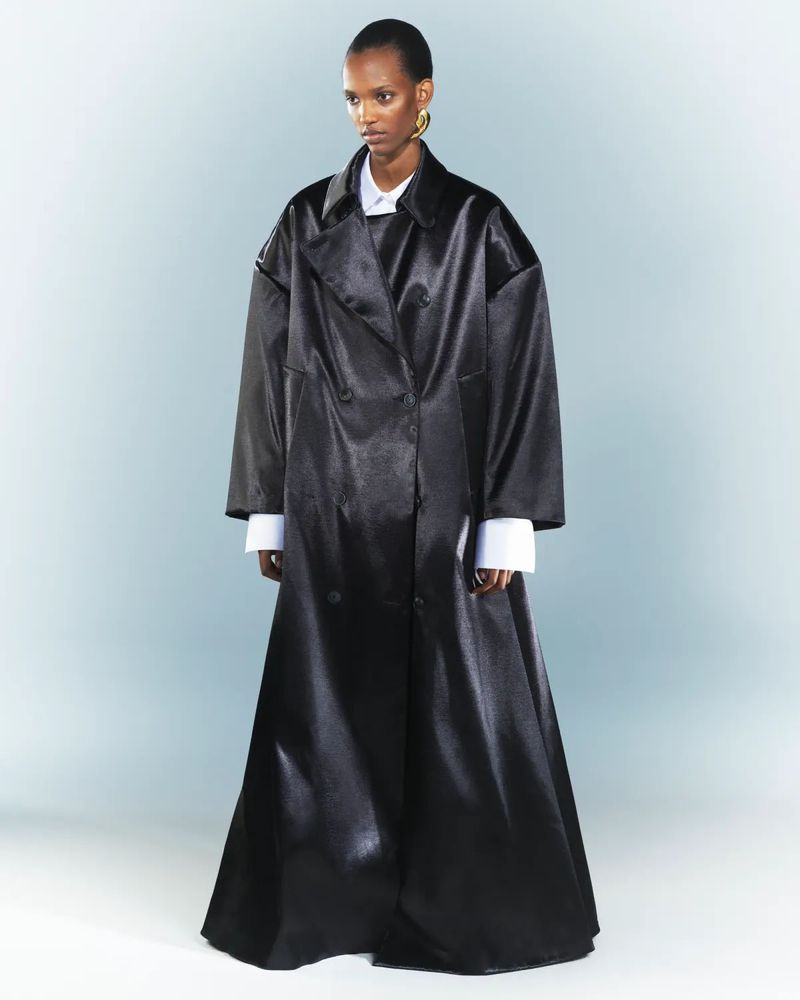 A black female fashion model posing in a floor length black coat with a white collared shirt underneath, accessorized with gold earrings.