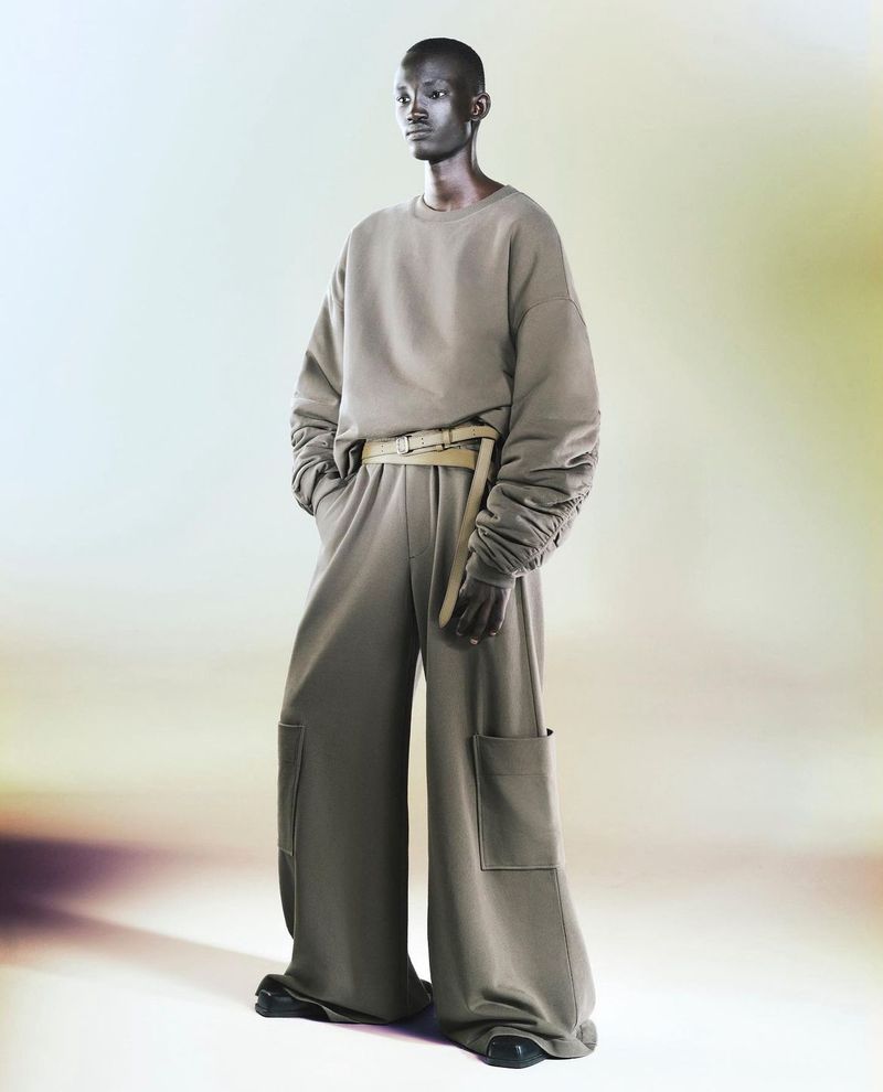 An adult fashion model poses in a photo studio wearing a sand-colored, oversized sweatshirt and wide-legged pants with a belt.