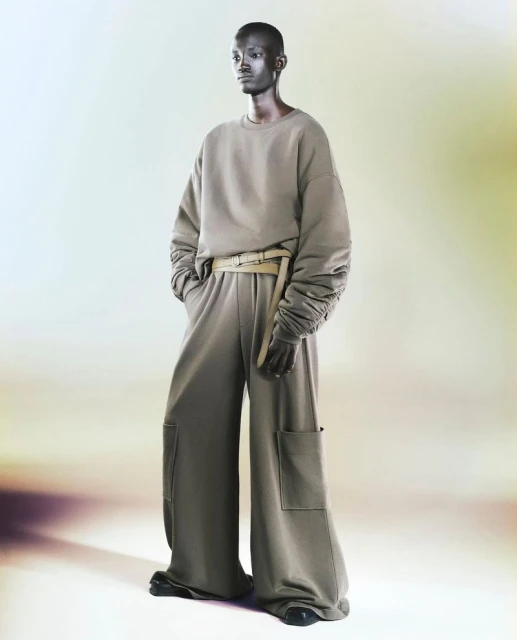 An adult fashion model poses in a photo studio wearing a sand-colored, oversized sweatshirt and wide-legged pants with a belt.