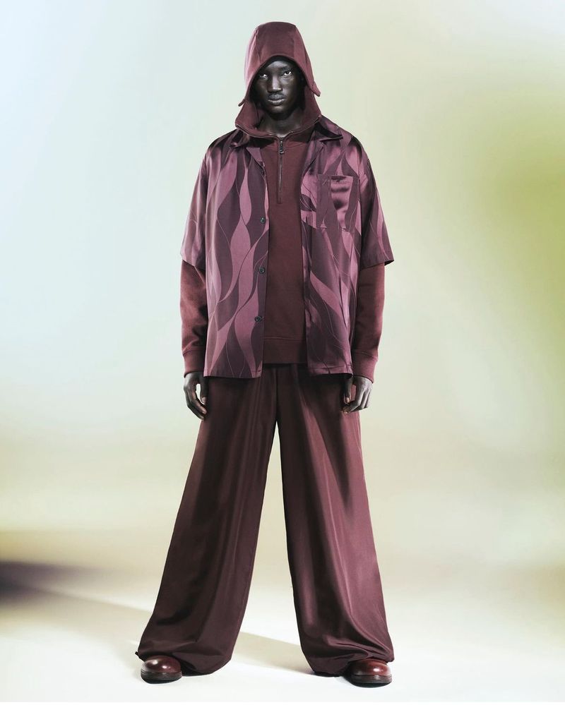 Full-length studio shot of a dark-skinned adult male wearing a hooded top, patterned shirt and wide-legged pants, posing on a gradient backdrop.