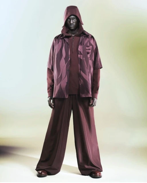 Full-length studio shot of a dark-skinned adult male wearing a hooded top, patterned shirt and wide-legged pants, posing on a gradient backdrop.