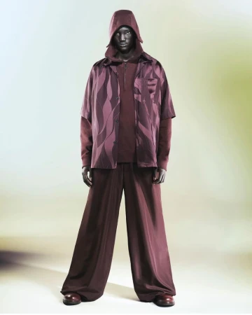 Full-length studio shot of a dark-skinned adult male wearing a hooded top, patterned shirt and wide-legged pants, posing on a gradient backdrop.