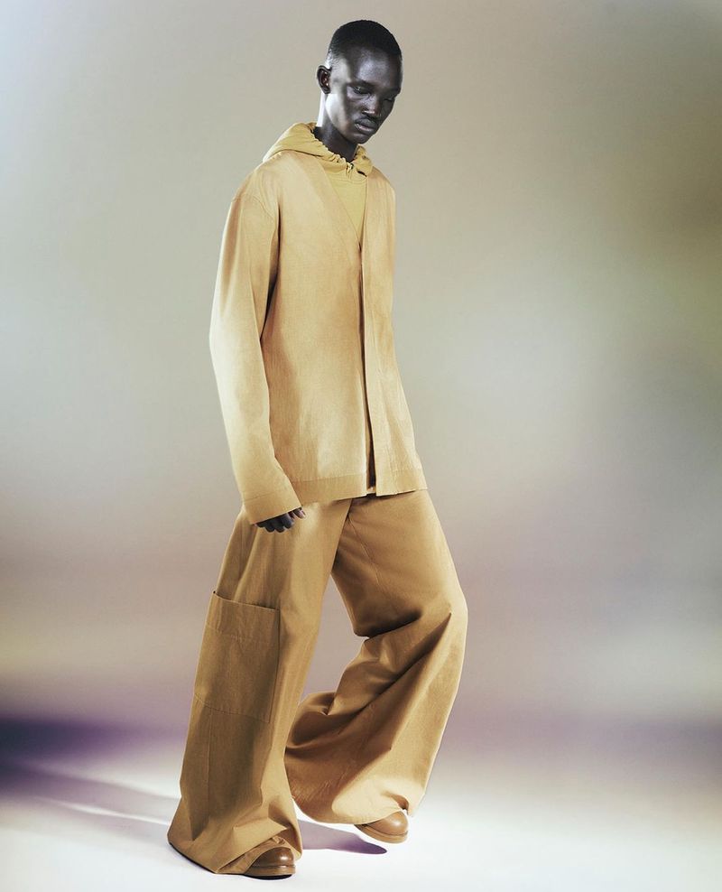 Dark-skinned male fashion model in a tan hooded jacket and matching wide-leg pants poses in a studio with an eye-level, full shot.