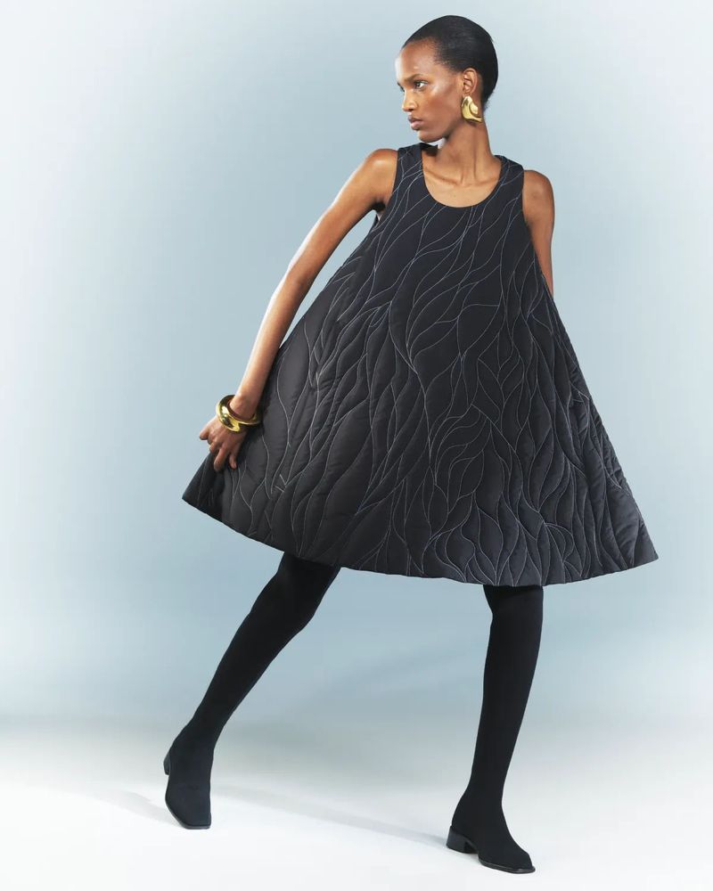 A woman with dark skin poses in a black quilted, patterned A-line dress, gold jewelry, and black tights and shoes against a light blue background.