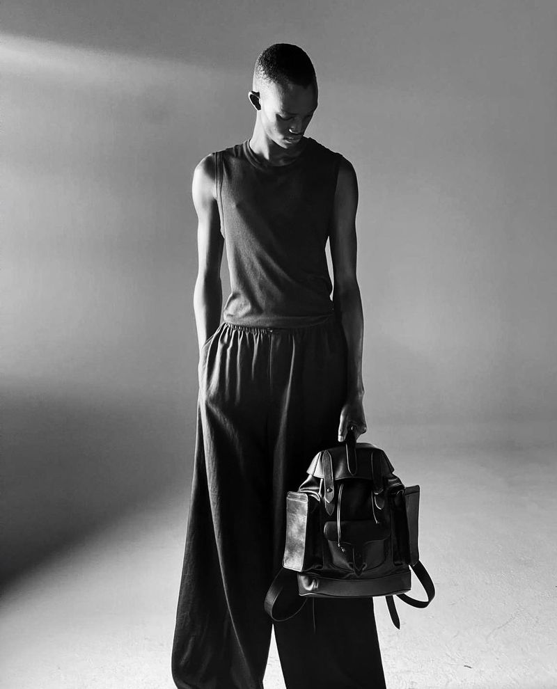 Monochromatic fashion shot of a slender person holding a black leather backpack in a photo studio, against a grey backdrop.