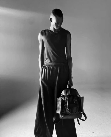 Monochromatic fashion shot of a slender person holding a black leather backpack in a photo studio, against a grey backdrop.
