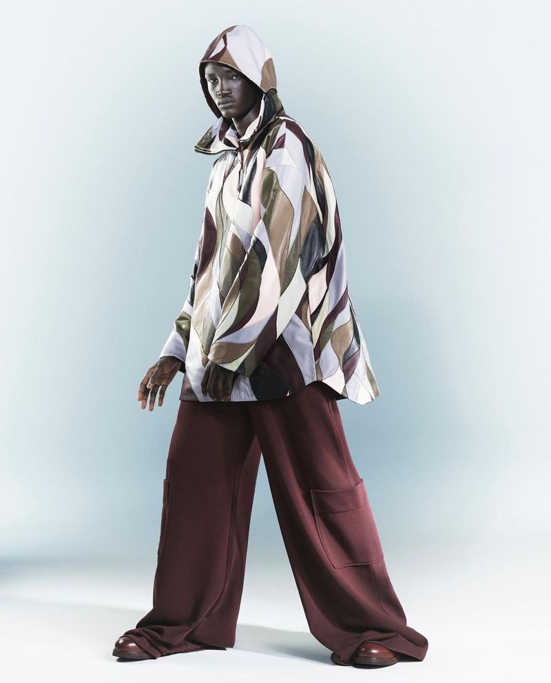 Fashion shot of a slender, dark-skinned man wearing a geometric-patterned hooded jacket and maroon wide-leg pants, posing in a studio.