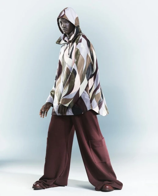 Fashion shot of a slender, dark-skinned man wearing a geometric-patterned hooded jacket and maroon wide-leg pants, posing in a studio.
