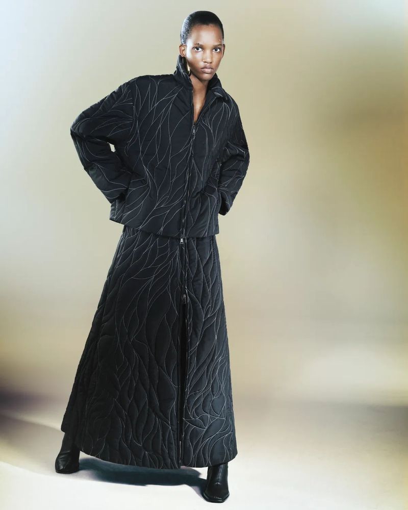 Fashion model poses in a studio wearing a black, quilted, zip-up jacket and long skirt with black heeled boots against a gradient background.