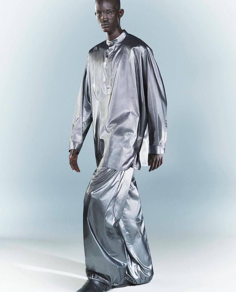 A full-length portrait of a Black male model in a shiny silver, long-sleeved tunic and draped skirt, standing against a gradient grey backdrop.
