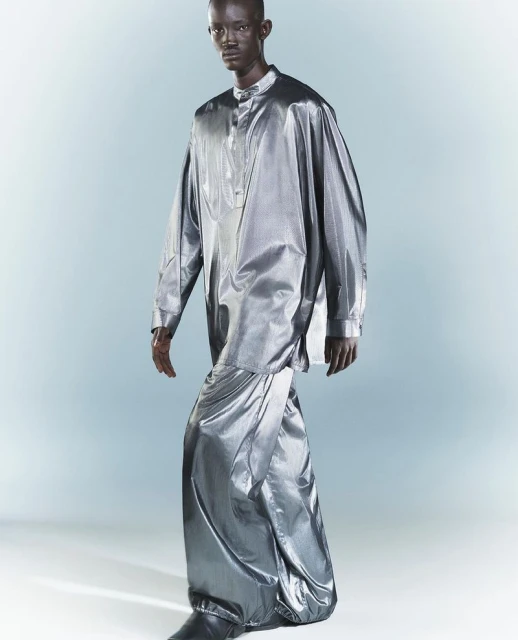 A full-length portrait of a Black male model in a shiny silver, long-sleeved tunic and draped skirt, standing against a gradient grey backdrop.