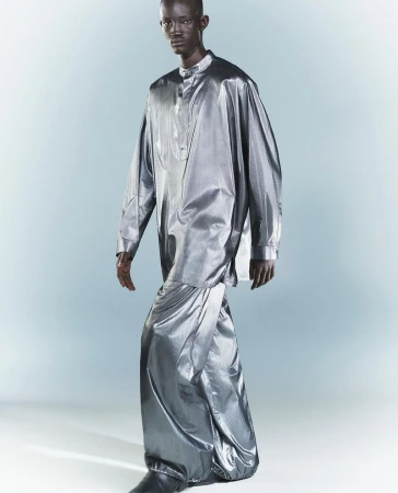 A full-length portrait of a Black male model in a shiny silver, long-sleeved tunic and draped skirt, standing against a gradient grey backdrop.