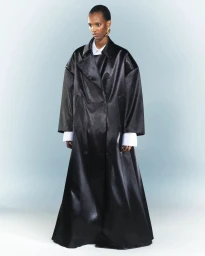 Edna Karibwami wearing an oversized floor-length black coat over a white collared shirt against a pale studio backdrop.