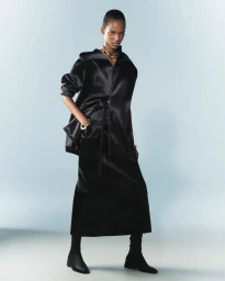 Model posing in an oversized black satin zip jacket, matching maxi skirt, gold jewelry, and black boots against a studio backdrop.