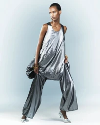Model wearing a metallic silver sleeveless draped tunic and matching wide-leg trousers with silver heels on a pale blue backdrop.