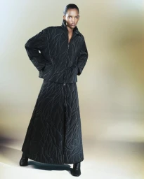 Model wearing a black quilted jacket and matching maxi skirt with contrast wavy stitching in a studio portrait.