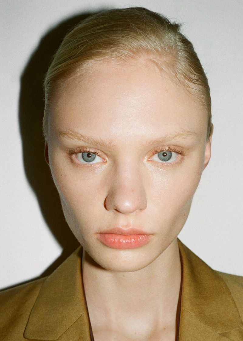 Close-up of a young adult female fashion model with blond hair and blue eyes wearing a tan suit jacket against a white backdrop.
