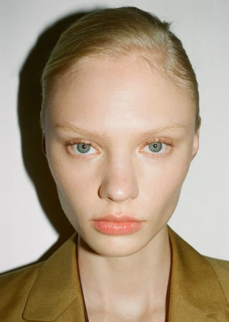 Close-up of a young adult female fashion model with blond hair and blue eyes wearing a tan suit jacket against a white backdrop.