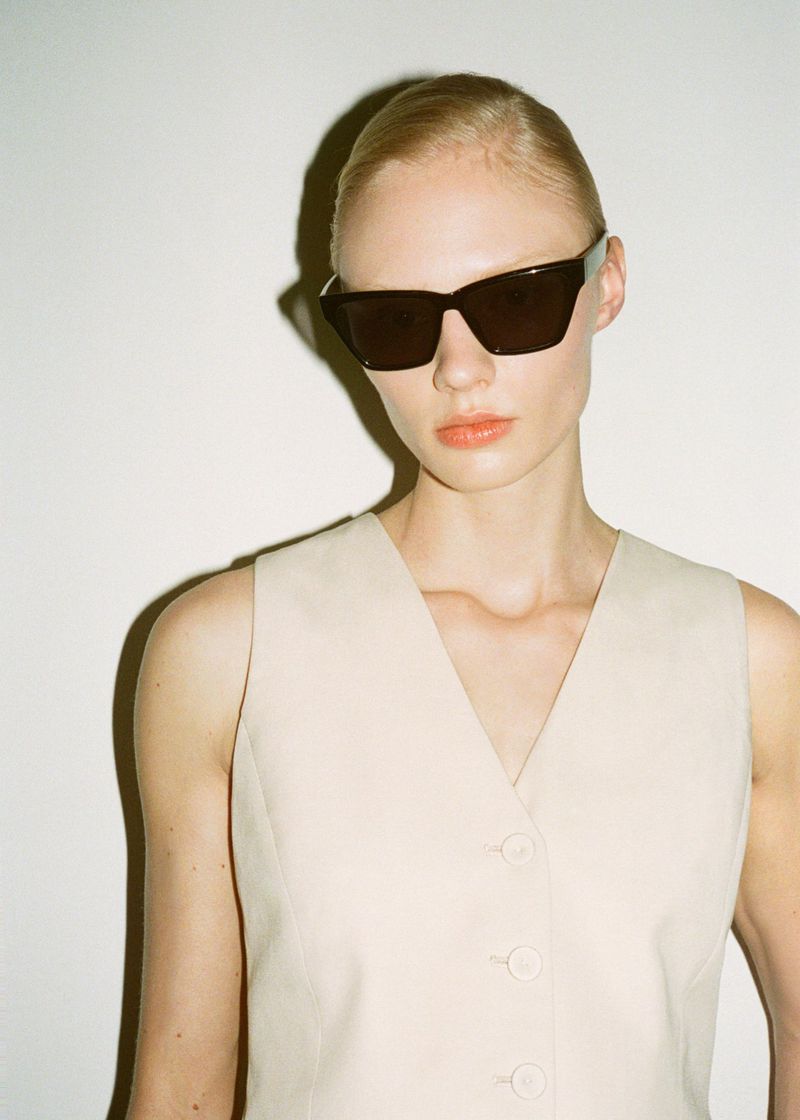 Portrait of a fair-skinned woman wearing dark sunglasses and a light beige button-up vest against a stark white background in a studio.