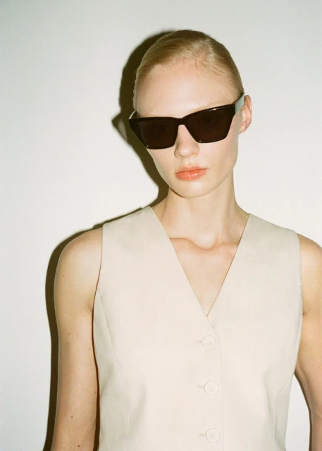 Portrait of a fair-skinned woman wearing dark sunglasses and a light beige button-up vest against a stark white background in a studio.