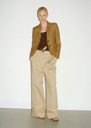 Model Vilma Sjöberg wearing a camel blazer and wide-leg beige trousers with a neck pouch against a white studio backdrop.