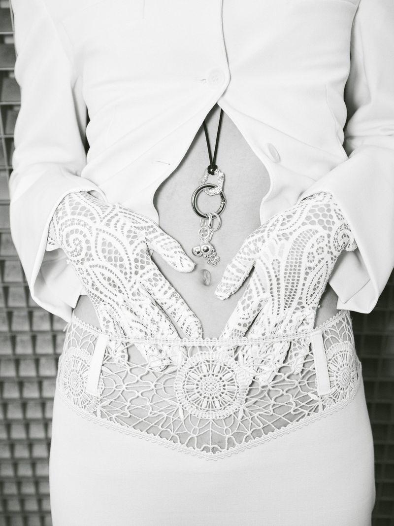 Close-up on a person's torso wearing a white jacket, lace gloves, and decorative lingerie, posing against a textured gray background.