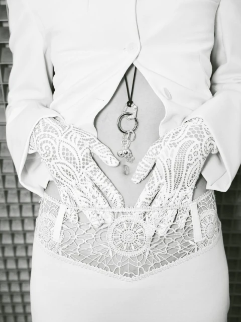 Close-up on a person's torso wearing a white jacket, lace gloves, and decorative lingerie, posing against a textured gray background.