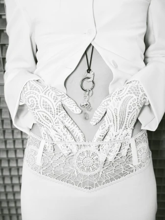 Close-up on a person's torso wearing a white jacket, lace gloves, and decorative lingerie, posing against a textured gray background.