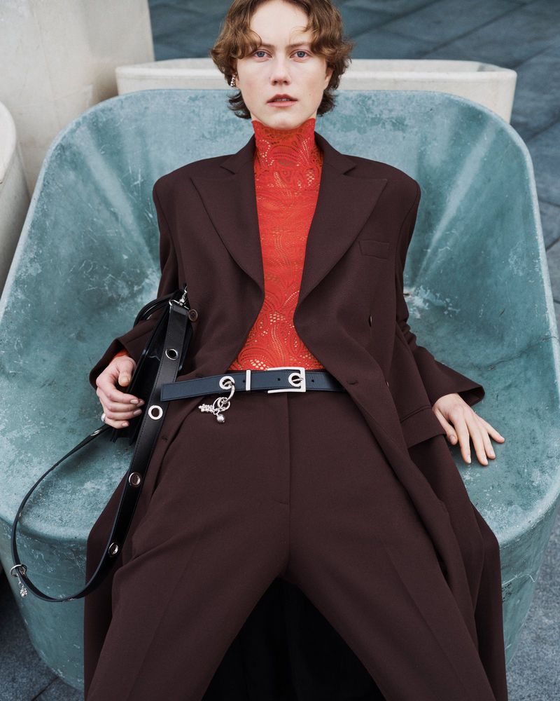 An adult woman with short brown hair sits in an aqua-colored chair wearing a brown pantsuit with a red lace top and a black shoulder bag.