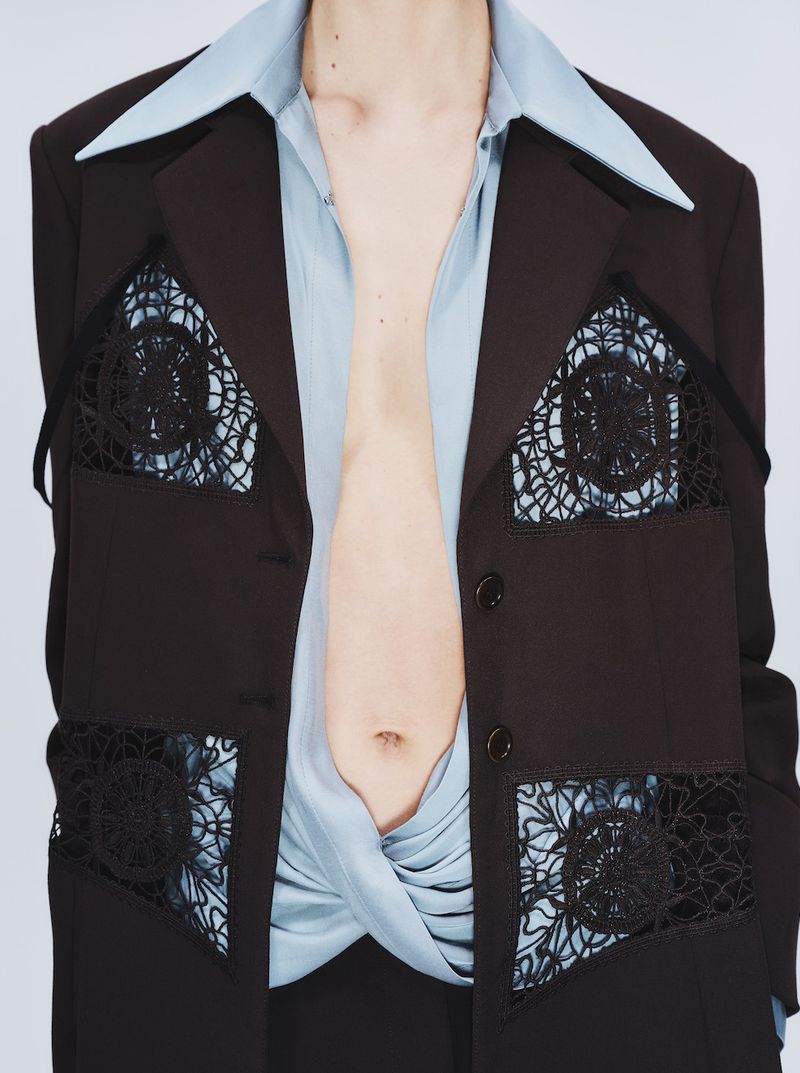 Model wearing a dark blazer with black lace cutout inserts over a light blue collared shirt against a white background