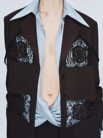 Model wearing a dark blazer with black lace cutout inserts over a light blue collared shirt against a white background