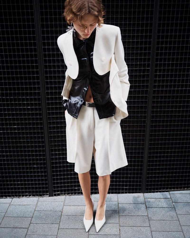 Fashion photo of a stylish woman wearing a white blazer, black shirt, white shorts, and heels, posing against a black fence and grey pavement.