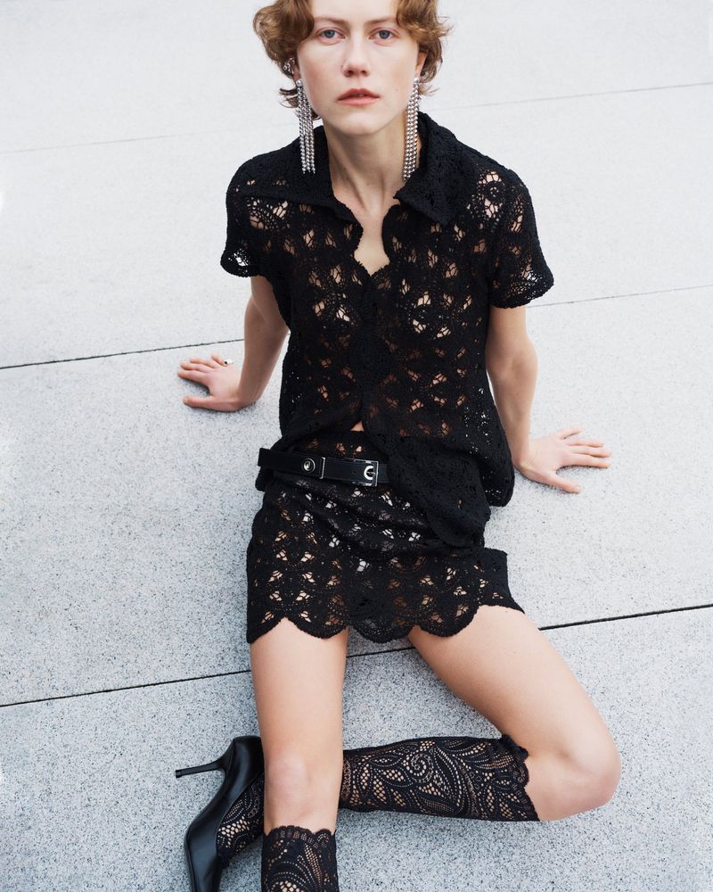 A fair-skinned young adult female sits on a grey surface wearing a black lace top, matching miniskirt with boots and long chandelier earrings.