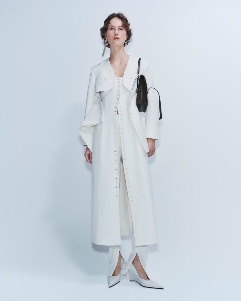 Woman wearing a long white coat, white shoes, and carrying a black handbag, standing in a photo studio with a white background.