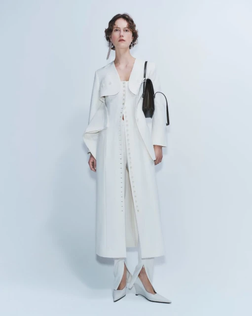 Woman wearing a long white coat, white shoes, and carrying a black handbag, standing in a photo studio with a white background.