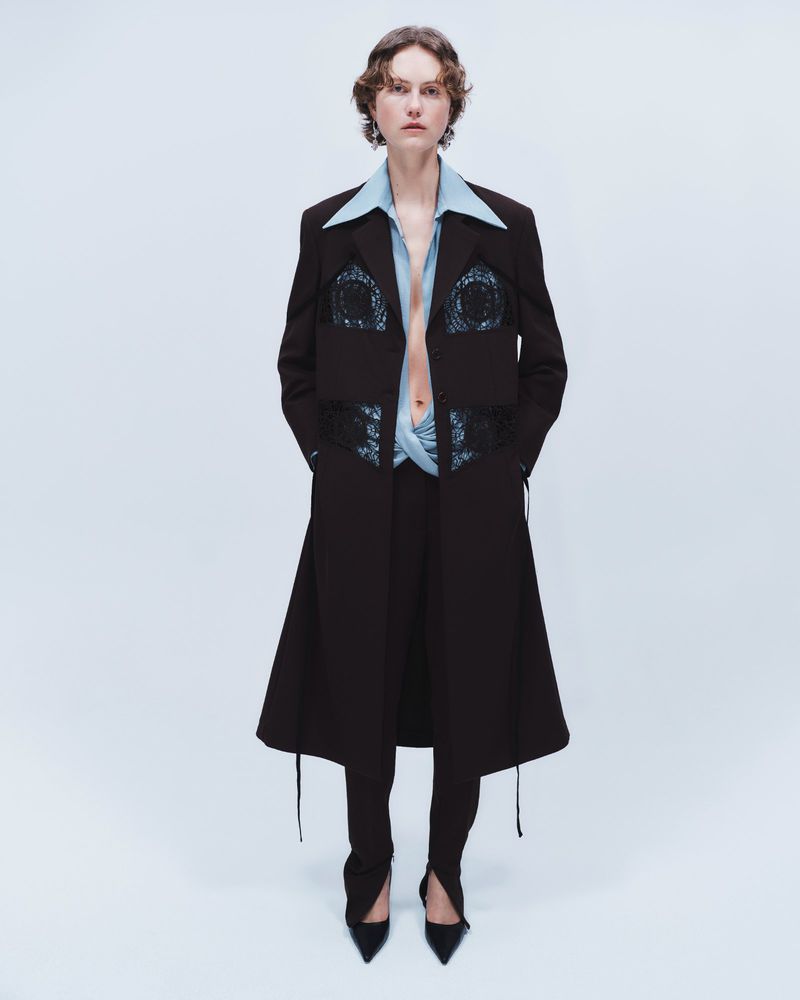 Julie Hoomans wearing a black tailored long coat and light blue collared shirt for Beaufille Pre Fall 2024 against a pale backdrop.